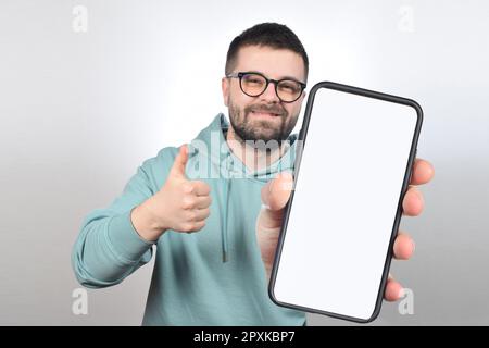 Ein junger Mann mit Brille und Bart zeigt ein Telefon mit einem leeren weißen Bildschirm Stockfoto
