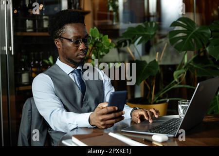 Junger afroamerikanischer Geschäftsmann, der ein Mobiltelefon mit einem Laptop verbindet und online arbeitet, während er im Café am Tisch sitzt Stockfoto