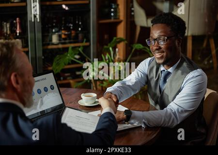 Geschäftspartner schütteln sich nach Verhandlungen die Hand, während sie während des Treffens im Restaurant am Tisch sitzen Stockfoto