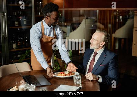 Ein afroamerikanischer Kellner serviert Geschäftsleuten mittags im Restaurant eine Mahlzeit Stockfoto