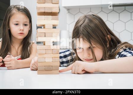 Zwei Schulmädchen und Kinder spielen Brettspiel Jenga auf dem Tisch in der Küche zu Hause, Bauturm aus Holzblöcken. Konzentriertes Mädchen versucht es Stockfoto