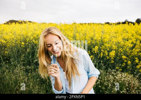 Wunderschöne, junge blonde, fröhliche Frau, die Löwenzahn in einem Feld blühender gelber Rapsblumen bläst Stockfoto