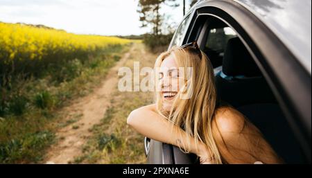 Eine junge lächelnde Frau sitzt im Auto und lehnt sich während ihres Roadtrips an das Fenster und genießt den wunderschönen Frühlingstag in der Natur Stockfoto