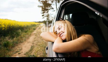Eine junge lächelnde Frau sitzt im Auto und lehnt sich während ihres Roadtrips an das Fenster und genießt den wunderschönen Frühlingstag in der Natur Stockfoto