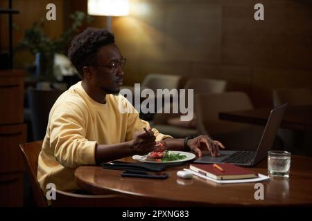 Afroamerikanischer Geschäftsmann, der online an einem Laptop arbeitet, am Tisch sitzt und im Café zu Mittag isst Stockfoto