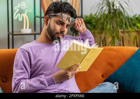 Ein junger indischer Mann liest ein Interessantes Buch, lächelt um und genießt Literatur, ruht sich auf einer bequemen Couch aus. Porträt eines friedlichen, fröhlichen hindu-Typen im Wohnraum auf dem Sofa Stockfoto