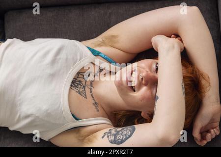 Eine Frau mit behaarten Achselhöhlen, die auf einem Sofa liegt, mit den Armen über dem Kopf. Stockfoto