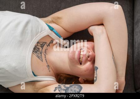 Eine Frau mit behaarten Achselhöhlen, die auf einem Sofa liegt, mit den Armen über dem Kopf. Stockfoto