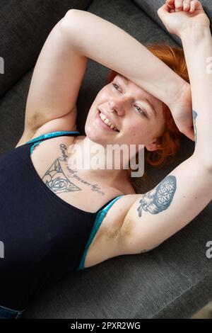 Eine Frau mit behaarten Achselhöhlen, die auf einem Sofa liegt, mit den Armen über dem Kopf. Stockfoto