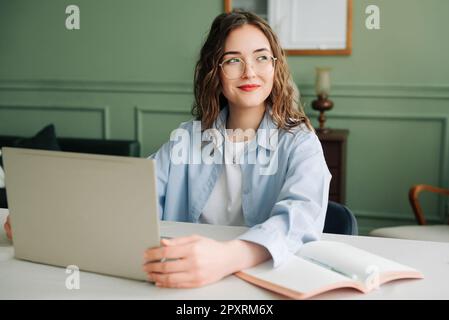 Lächelnde weibliche Mitarbeiterin, die in einem modernen Büro mit einem Notebook arbeitet, Marketingmanagerin oder Geschäftsfrau, die sich auf den Bildschirm konzentrierte, glücklich und selbstbewusst Stockfoto