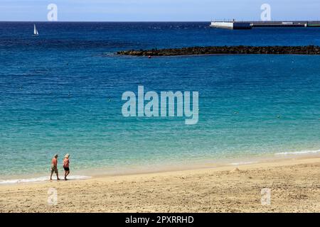 Ein älteres Ehepaar blickt aufs Meer und spaziert entlang eines wunderschönen Sandstrandes mit tiefblauem Meer und einem Segelboot in der Ferne. Lanzarote Stockfoto