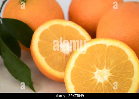 Nahaufnahme eines Orangenspalts in zwei Teilen neben anderen ganzen Orangen mit einigen Blättern des Baumes, die noch frisch auf neutralem Hintergrund sind Stockfoto