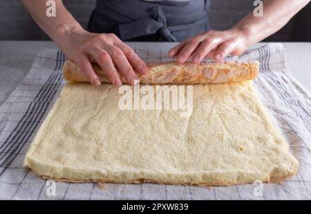 Frisch gebackener Biskuitkuchen für schweizer Brötchen, zum Abkühlen aufgerollt auf einem zuckerhaltigen Geschirrtuch von Frauenhänden Stockfoto
