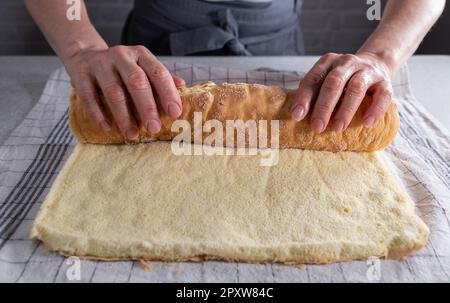 Frisch gebackener Biskuitkuchen für schweizer Brötchen, zum Abkühlen aufgerollt auf einem zuckerhaltigen Geschirrtuch von Frauenhänden Stockfoto
