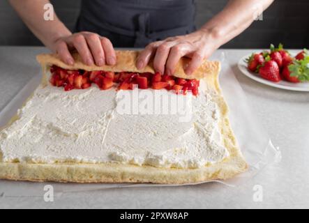 schweizer Brötchen. Mit Schlagsahne und Erdbeerfüllung von Frauenhänden auf dem Küchentisch zusammengerollt. Stockfoto