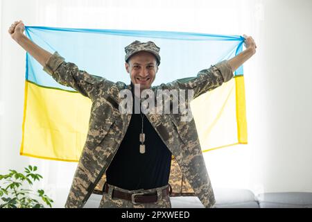 Ukrainischer Mann-Krieger, der mit einer Militär-Pixel-Uniform bekleidet ist, hält die gelb-blaue Flagge des Staates Ukraine und an der Kette ein kleines Wappen von Stockfoto