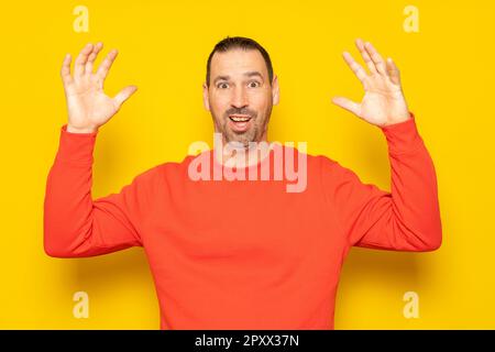 Ein bärtiger hispanischer Mann in seinen 40s Jahren, der einen roten Pullover trägt, der von einem unerwarteten Besucher unglaublich überrascht wurde, hebt seine Arme aufgeregt. Isoliert auf gelb Stockfoto