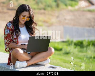 Eine junge Frau mit Sonnenbrille, die einen Laptop benutzt, während sie auf einer Bank in einem Park sitzt. Technologiekonzept. Stockfoto