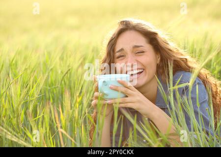 Glückliche Frau, die lacht, Kaffee auf einem Feld trinkt und in die Kamera schaut Stockfoto