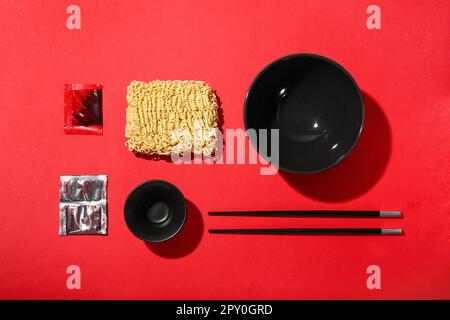 Rohe Instantnudeln mit leeren Schüsseln, Stäbchen und Gewürzen auf rotem Hintergrund Stockfoto