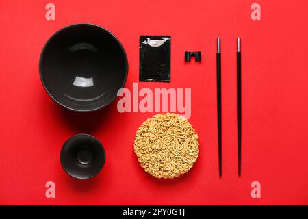 Rohe Instantnudeln mit leeren Schüsseln, Stäbchen und Gewürzen auf rotem Hintergrund Stockfoto