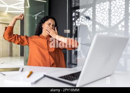 Müde Geschäftsfrau im Büro, die sich streckt und gähnt, lateinamerikanische Frau, die im Innenbereich am Arbeitsplatz arbeitet, mit einem gelangweilten Laptop übermüdet. Stockfoto