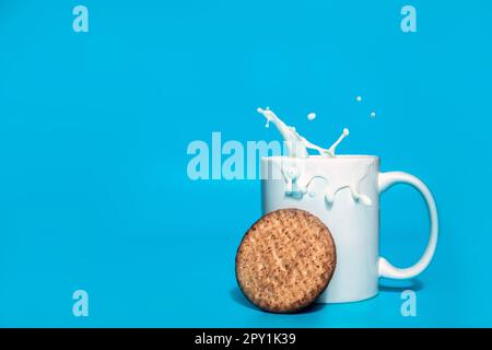 Haferflocken-Kekse fallen in eine weiße Tasse mit Milch und einem Spritzer auf blauem Hintergrund mit Kopierraum, normale gesunde Getreidekekse, Milchgetränkprodukt, hea Stockfoto