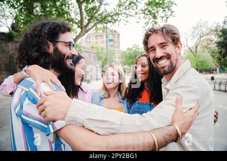 Gruppe von jungen erwachsenen, multiethnischen Menschen, die Spaß miteinander haben, sich gegenseitig zu umarmen. Ein glücklicher Mann, der in die Kamera schaut und seine Freunde im Kreis umarmt. Union eines fröhlichen und positiven Teams. Hochwertiges Foto Stockfoto