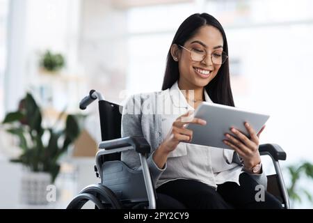 Glücklich und erfolgreich. Eine attraktive junge Geschäftsfrau im Rollstuhl, die ihr Tablet im Büro benutzt. Stockfoto