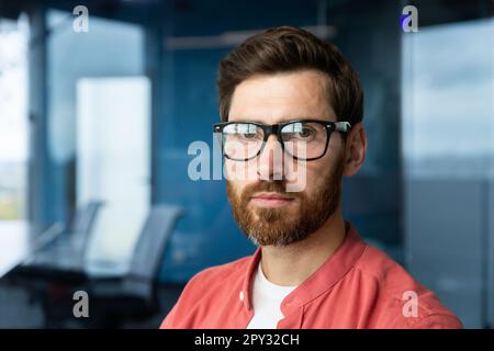 Nahaufnahme. Porträt eines jungen Profis, eines Brillenentwicklers und eines roten Hemds, das selbstbewusst in die Kamera schaut. Stockfoto