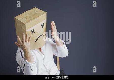 Ein trauriger Mann in einem weißen Hemd mit einer Tasche auf dem Kopf, mit einem gezogenen traurigen Emoticon hat Angst, hält seine Hände nahe seinem Gesicht. Emotionen und Gesten. Stockfoto