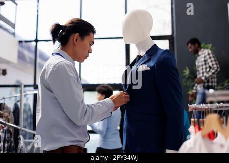 Asiatischer Arbeiter arrangiert Schaufensterpuppe mit formellem Anzug, arbeitet im Laden visuell mit modischer Ware. Mitarbeiter bereitet die Eröffnung der Boutique vor und wartet im Einkaufszentrum auf Kunden Stockfoto