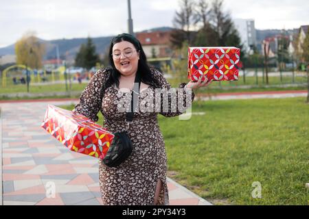 Eine schöne, glückliche junge Frau in Übergröße mit zwei Geschenken, die in einem Park herumläuft Stockfoto