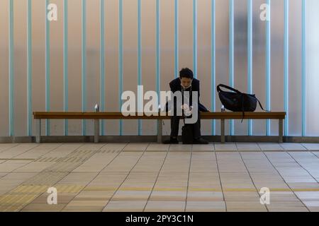 Ein japanischer Büroangestellter oder Angestellter sitzt auf einer Bank, um sein Smartphone in Futako-Shinchi, Station, Tokio, Japan, zu verwenden. Stockfoto