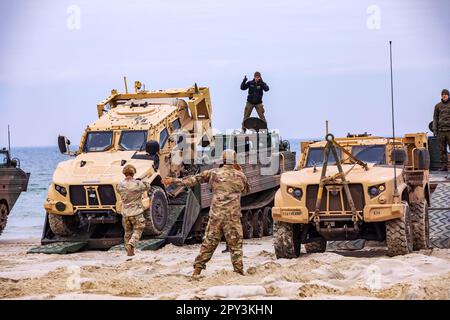 Bemowo Piskie, Polen. 17. April 2023. Soldaten, die dem 2. Panzerbrigade-Kampfteam, der 1. Kavallerieabteilung, der NATO EFP-Gefechtsgruppe Polen, zur Unterstützung der 4. Infanteriedivision zugewiesen wurden, zusammen mit polnischen Soldaten, die der 15. Mechanisierten Infanteriedivision zugeteilt wurden, während der Übung Zalew 23 gemeinsame leichte taktische Fahrzeuge auf mittelgroße Amphibientransporte laden; Eine amphibische Angriffsübung in Danzig, Polen, 17. April 2023. Die Mission der 4. Infanterieabteilung in Europa besteht darin, sich an multinationalen Ausbildungsmaßnahmen und Übungen auf dem ganzen Kontinent zu beteiligen, die mit NATO-Verbündeten und der regionalen Sicherheit zusammenarbeiten Stockfoto