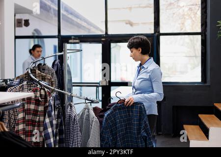 Der stilvolle Manager bereitet den Laden auf die Eröffnung vor und arrangiert elegante Kleidung an Kleiderbügeln im Einkaufszentrum. Eine weiße Frau, die sich modische Ware anschaut, den Preis in einer modernen Boutique überprüft Stockfoto
