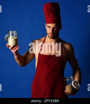 Ein seriöser Koch mit burgunderfarbenem Hut und Schürze hält Glasgefäße mit Grobgrieß und Müsli. Professioneller Koch mit Kochhut. Frühstück und gesundes Essen. Diät Stockfoto