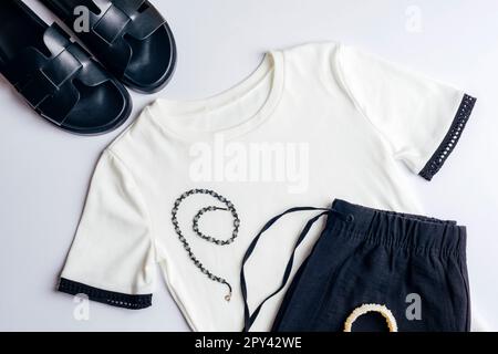 Weißes T-Shirt, schwarze Shorts oder Hosen, schwarze Hausschuhe, Halskette und Armband. Blick von oben auf die Freizeitkleidung der Frau. Trendiger Hipster-Look. Flach Stockfoto