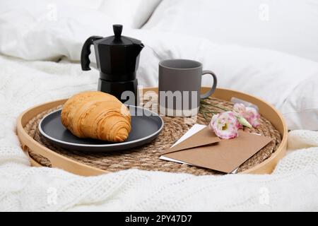 Tablett mit leckeren Croissants, einer Tasse Kaffee und Blumen auf weißem Bett Stockfoto