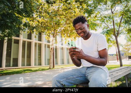 Ein glücklicher afroamerikanischer junger Mann, der sein Handy am College benutzt hat, schickt Textnachrichten draußen auf einer Bank. Porträt eines Schülers, der auf dem Campus auf den Bildschirm seines Mobiltelefons schaut. Hochwertiges Foto Stockfoto