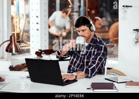 Kaffee hält mich an meinen arbeitsreichen Tagen am Laufen. Ein gutaussehender junger Geschäftsmann, der Kaffee trinkt, während er in einem Büro in seiner Werkstatt an einem Laptop arbeitet. Stockfoto