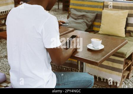 afroamerikanischer Mann sitzt am Tisch im Café mit einer Tasse Kaffee auf der Sommerveranda, hält ein Smartphone und gibt die Kreditkartennummer ein. Rückansicht Stockfoto