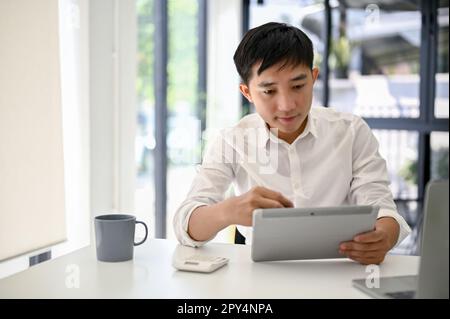 Intelligenter und professioneller asiatischer Geschäftsmann oder männlicher Chef, der sein Tablet verwendet und im Büro arbeitet. Stockfoto