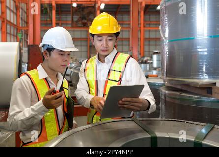 Junger asiatischer Ingenieurleiter schult neue Mitarbeiter in der Blechfabrik. Alle tragen Schutzweste und Schutzhelm. Stahlblechrollenstapel ist eingefahren Stockfoto