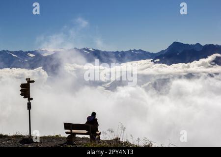 Ein Wanderer am Ufer beobachtet, wie der Nebel zwischen den Bergen aufgeht und die alpengipfel im Hintergrund zu sehen sind Stockfoto