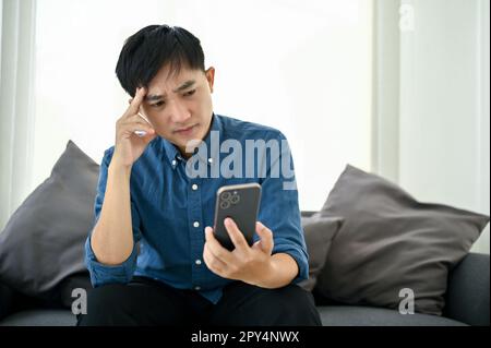 Gestresster und aufmerksamer asiatischer Millennials, der auf sein Smartphone schaut, etwas auf dem Bildschirm liest, mit ernsthaftem Gesicht, während er auf dem Sofa sitzt Stockfoto