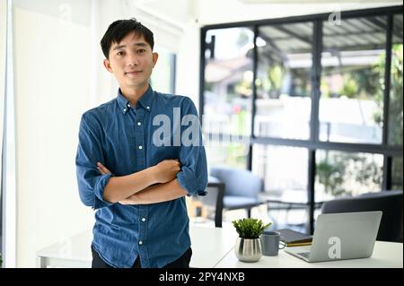 Kluge und glückliche männliche Büroangestellte oder Programmierer in lässiger Kleidung stehen in seinem Büro mit verschränkten Armen. Stockfoto