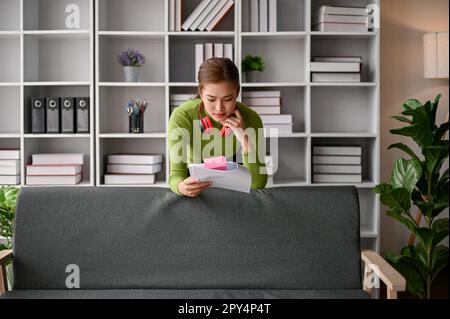 Fokussierte und seriöse asiatische Geschäftsfrau in lässiger Kleidung, die sich in ihrem Büro auf ein Sofa lehnt, Dokumente liest und einen Lösungsplan findet. Stockfoto