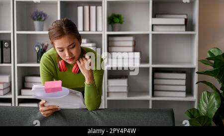 Fokussierte asiatische Geschäftsfrau in lässiger Kleidung, die sich in ihrem Büro auf ein Sofa lehnt und Dokumente liest. Stockfoto