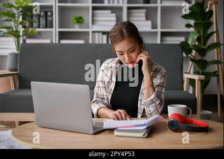 Fokussierte junge asiatische Freiberuflerin liest Dokumente und arbeitet an ihrem Auftrag an einem Kaffeetisch in ihrem Wohnzimmer. Stockfoto